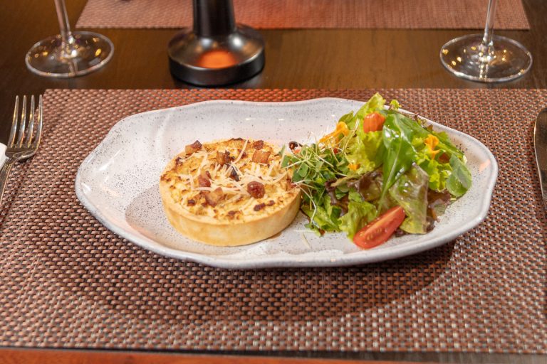 Quiche Loraine: Clásica tarta salada de origen francés, masa quebrada con relleno de panceta crocante y queso ricota, acompañada de mix de hojas frescas y tomates cherrys.